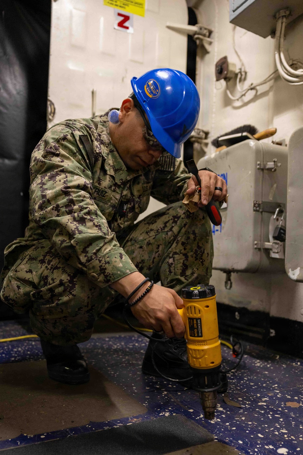 A U.S. Navy sailor identified by the name tape 'RUIZ' is crouched down on the deck of what appears to be a naval vessel, operating a yellow heat gun tool. The sailor is wearing Navy Working Uniform (NWU) camouflage, a blue hard hat with a Navy emblem, safety glasses, and ear protection. The work appears to involve applying heat to flooring material, likely for installation or removal purposes.