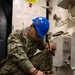 A U.S. Navy sailor identified by the name tape 'RUIZ' is crouched down on the deck of what appears to be a naval vessel, operating a yellow heat gun tool. The sailor is wearing Navy Working Uniform (NWU) camouflage, a blue hard hat with a Navy emblem, safety glasses, and ear protection. The work appears to involve applying heat to flooring material, likely for installation or removal purposes.