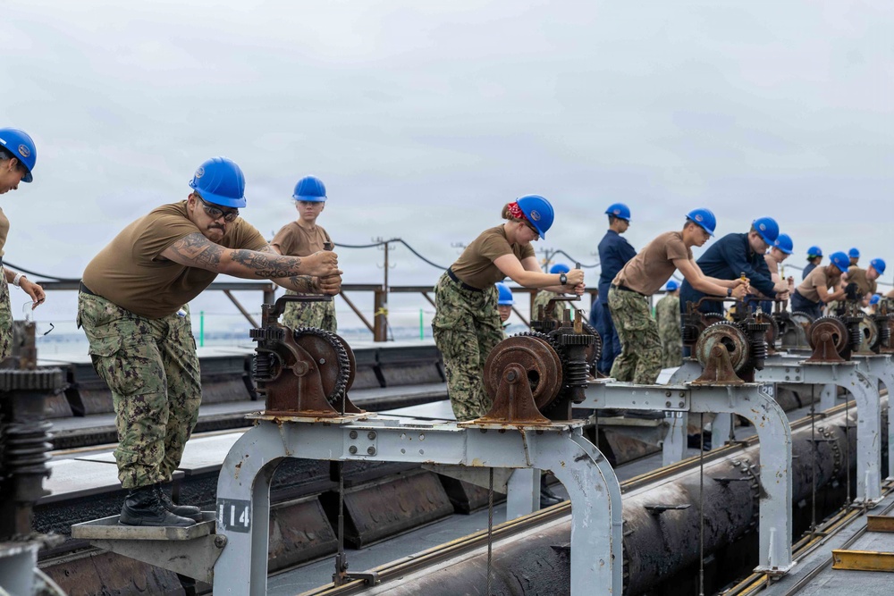 A group of U.S. Navy personnel wearing blue hard hats and camouflage uniforms are working in a coordinated line on what appears to be the deck of a naval vessel or dry dock structure. They are manually operating gear-driven mechanical mechanisms mounted on metal frames, suggesting a synchronized operation such as opening or closing flood gates, hatches, or ballast controls. The overcast sky and industrial maritime setting indicate an outdoor naval facility or ship deck.