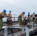 A group of U.S. Navy personnel wearing blue hard hats and camouflage uniforms are working in a coordinated line on what appears to be the deck of a naval vessel or dry dock structure. They are manually operating gear-driven mechanical mechanisms mounted on metal frames, suggesting a synchronized operation such as opening or closing flood gates, hatches, or ballast controls. The overcast sky and industrial maritime setting indicate an outdoor naval facility or ship deck.