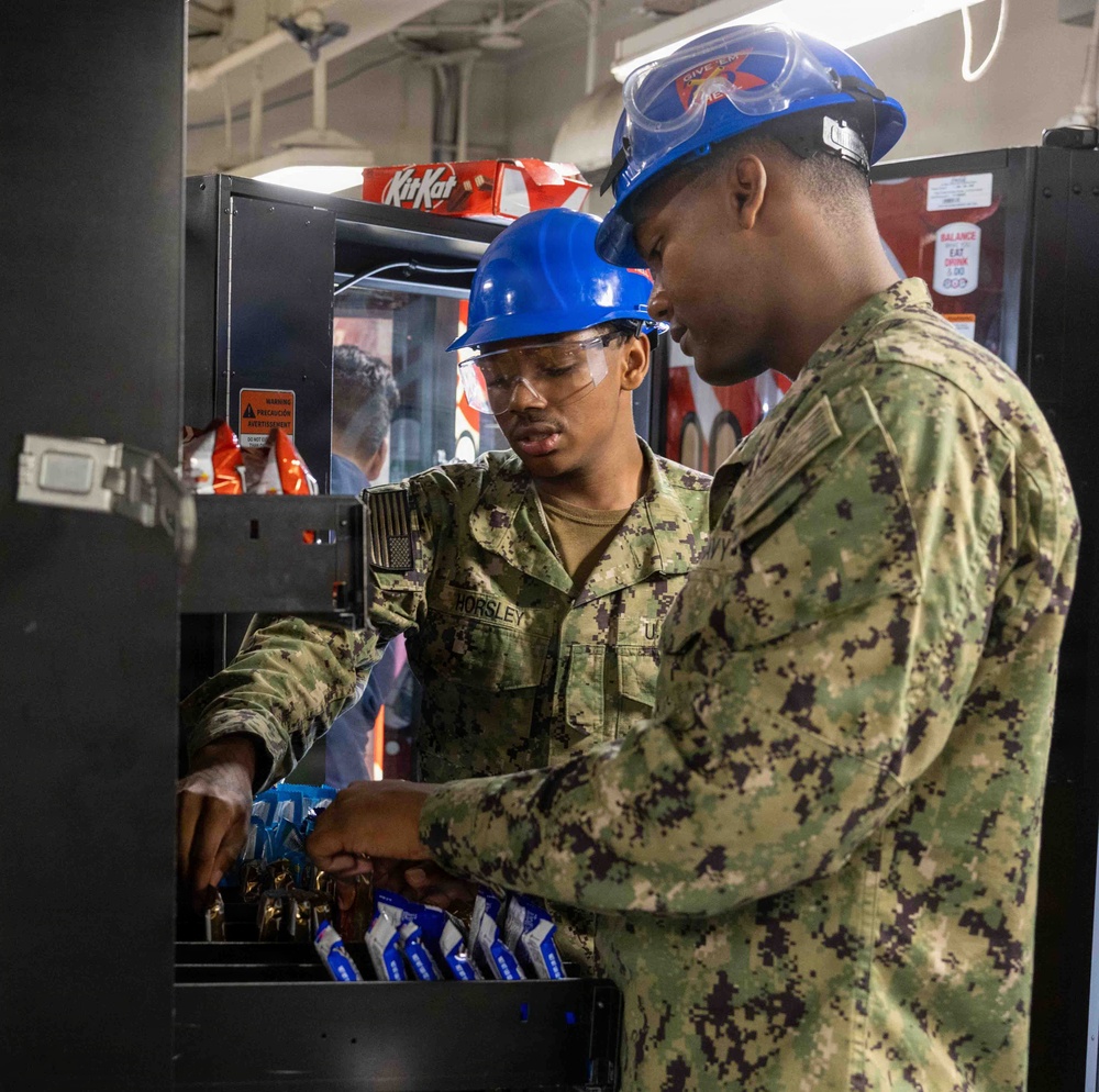 Two U.S. Navy sailors wearing blue hard hats and safety goggles are stocking or servicing a vending machine. Both are dressed in Navy Working Uniform (NWU) camouflage pattern, and one sailor's name tape reads 'HORSLEY'. They appear to be loading snack items into the machine in what looks like a shipboard or military facility environment.