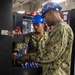 Two U.S. Navy sailors wearing blue hard hats and safety goggles are stocking or servicing a vending machine. Both are dressed in Navy Working Uniform (NWU) camouflage pattern, and one sailor's name tape reads 'HORSLEY'. They appear to be loading snack items into the machine in what looks like a shipboard or military facility environment.