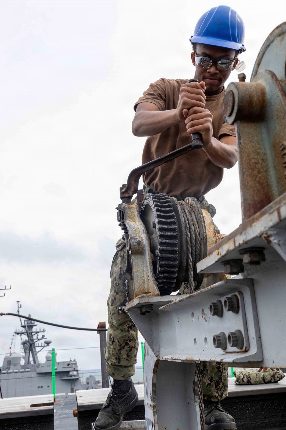 A U.S. Navy sailor in digital camouflage utilities and a blue hard hat operates a manual winch or capstan mechanism on what appears to be a naval vessel or pier. The sailor is using a breaker bar or ratchet handle to turn the gear mechanism, with steel cable wound around the drum. A naval warship is visible in the background, suggesting a port or harbor setting.