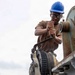 A U.S. Navy sailor in digital camouflage utilities and a blue hard hat operates a manual winch or capstan mechanism on what appears to be a naval vessel or pier. The sailor is using a breaker bar or ratchet handle to turn the gear mechanism, with steel cable wound around the drum. A naval warship is visible in the background, suggesting a port or harbor setting.