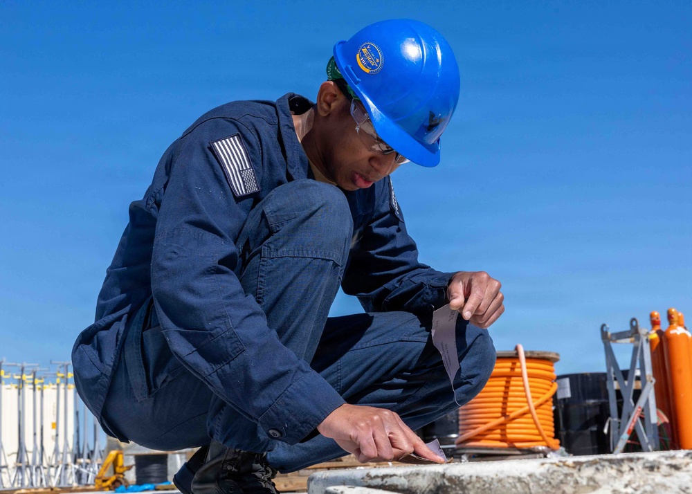 A worker in navy blue coveralls and a blue hard hat is crouching on what appears to be an outdoor industrial or naval surface, applying or handling tape/material on a flat surface. The individual is wearing safety glasses and has a subdued American flag patch visible on the left sleeve. The setting appears to be an outdoor work environment with equipment including an orange cable spool and gas cylinders visible in the background.