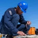 A worker in navy blue coveralls and a blue hard hat is crouching on what appears to be an outdoor industrial or naval surface, applying or handling tape/material on a flat surface. The individual is wearing safety glasses and has a subdued American flag patch visible on the left sleeve. The setting appears to be an outdoor work environment with equipment including an orange cable spool and gas cylinders visible in the background.