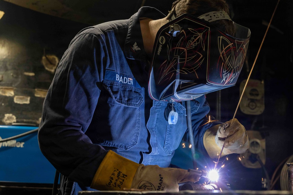 A welder named 'Baden' is actively welding metal in what appears to be a workshop or industrial setting. The welder is wearing a decorated Miller brand auto-darkening welding helmet and leather welding gloves, with visible sparks and arc light at the work point. The image captures a professional or vocational welding environment with Miller welding equipment visible in the background.