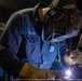 A welder named 'Baden' is actively welding metal in what appears to be a workshop or industrial setting. The welder is wearing a decorated Miller brand auto-darkening welding helmet and leather welding gloves, with visible sparks and arc light at the work point. The image captures a professional or vocational welding environment with Miller welding equipment visible in the background.