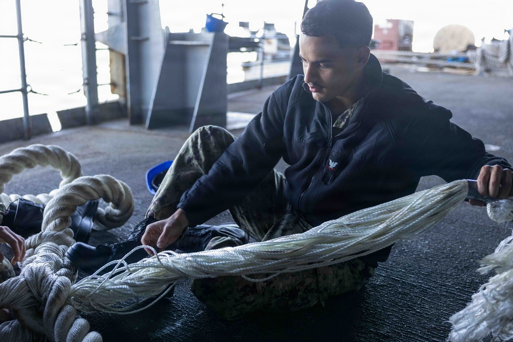 A young male sailor or military personnel is seated on a ship's deck working with large white mooring or dock lines. He is wearing a black jacket and camouflage pants, carefully handling and organizing the rope. The setting appears to be the interior deck area of a naval vessel with water visible in the background.