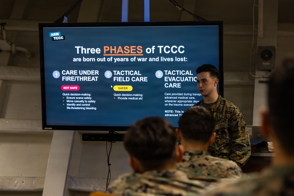 A military instructor in camouflage uniform is presenting a TCCC (Tactical Combat Casualty Care) training briefing to a group of uniformed personnel. The presentation slide displayed on a large monitor outlines the 'Three Phases of TCCC,' covering Care Under Fire/Threat, Tactical Field Care, and Tactical Evacuation Care. The setting appears to be an indoor military training facility or shipboard compartment.