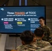 A military instructor in camouflage uniform is presenting a TCCC (Tactical Combat Casualty Care) training briefing to a group of uniformed personnel. The presentation slide displayed on a large monitor outlines the 'Three Phases of TCCC,' covering Care Under Fire/Threat, Tactical Field Care, and Tactical Evacuation Care. The setting appears to be an indoor military training facility or shipboard compartment.