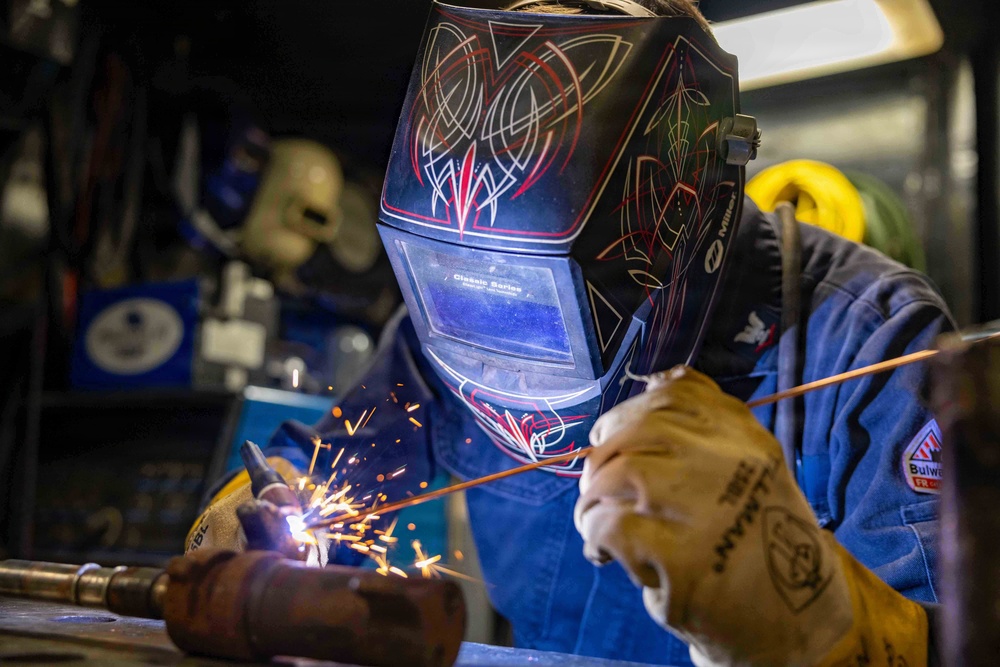 A welder is actively performing a welding operation in an industrial workshop setting. The worker is equipped with full protective gear including a decorative Miller 'Classic Series' auto-darkening welding helmet and Tillman leather welding gloves, along with a Bulwark FR (flame-resistant) blue work jacket. Sparks are visibly flying from the welding point, indicating active metal fusion work.