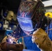 A welder is actively performing a welding operation in an industrial workshop setting. The worker is equipped with full protective gear including a decorative Miller 'Classic Series' auto-darkening welding helmet and Tillman leather welding gloves, along with a Bulwark FR (flame-resistant) blue work jacket. Sparks are visibly flying from the welding point, indicating active metal fusion work.