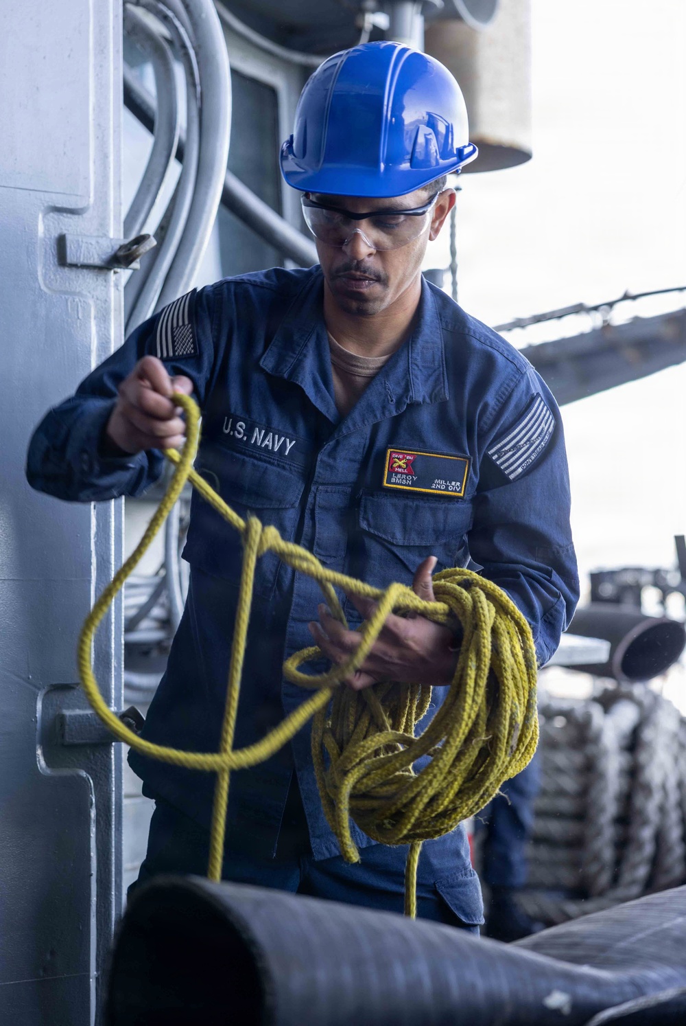 A U.S. Navy sailor wearing a blue hard hat and safety glasses is handling a coiled yellow rope on what appears to be the deck of a naval vessel. The sailor's uniform displays 'U.S. NAVY' on the chest along with name patches reading 'LEROY' and rank 'BMSN' (Boatswain's Mate Seaman), assigned to 'MILLER 2ND DIV'. The image captures a routine deck seamanship task aboard a warship.