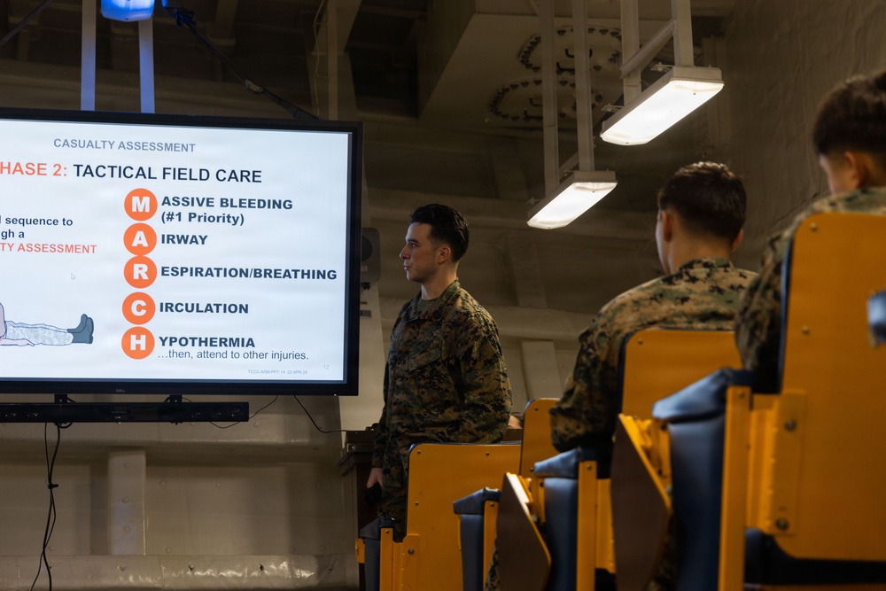 Military personnel in camouflage uniforms are attending a Tactical Combat Casualty Care (TCCC) training session. A large display screen shows a 'Casualty Assessment - Phase 2: Tactical Field Care' slide featuring the MARCH acronym protocol. The setting appears to be a briefing room or training facility aboard a military vessel or installation.