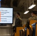 Military personnel in camouflage uniforms are attending a Tactical Combat Casualty Care (TCCC) training session. A large display screen shows a 'Casualty Assessment - Phase 2: Tactical Field Care' slide featuring the MARCH acronym protocol. The setting appears to be a briefing room or training facility aboard a military vessel or installation.