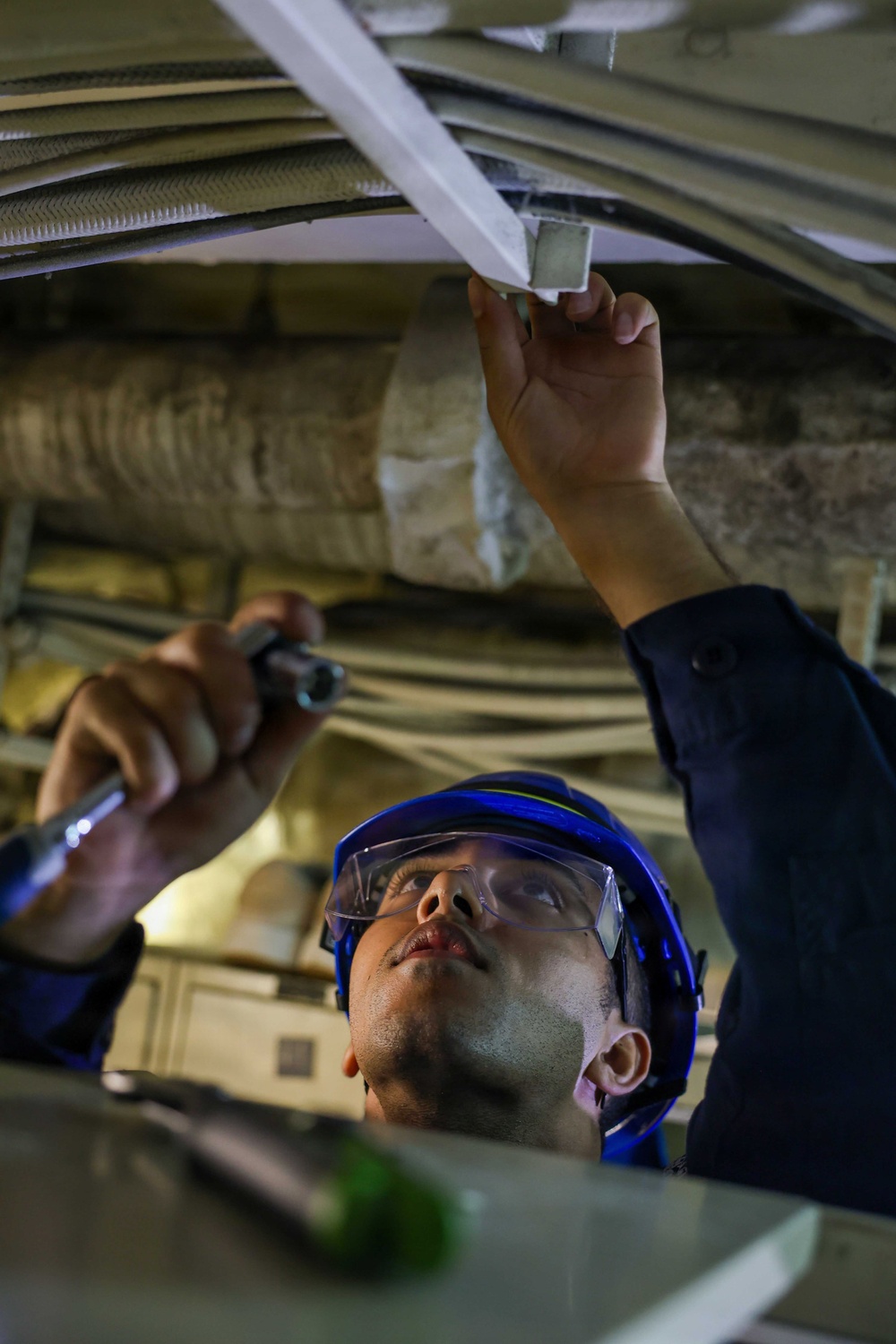 A technician wearing a blue hard hat and safety glasses is performing maintenance or inspection work in a confined overhead space. He is holding a flashlight in one hand while manipulating or examining a white structural component above him with the other hand. The environment appears to be an industrial or mechanical space with visible cables, insulated pipes, and structural framing.