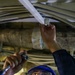 A technician wearing a blue hard hat and safety glasses is performing maintenance or inspection work in a confined overhead space. He is holding a flashlight in one hand while manipulating or examining a white structural component above him with the other hand. The environment appears to be an industrial or mechanical space with visible cables, insulated pipes, and structural framing.