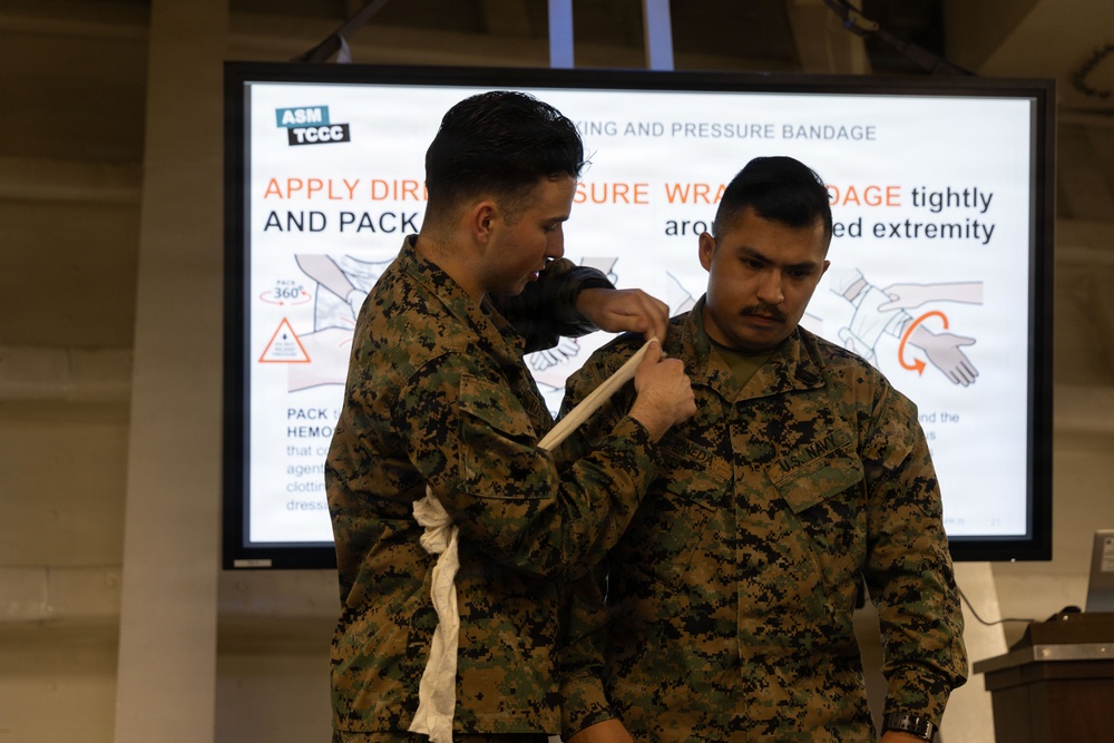 Two U.S. military personnel in MARPAT camouflage uniforms are conducting a Tactical Combat Casualty Care (TCCC) training demonstration. One service member is applying a pressure bandage or wound packing technique to the other's shoulder/upper arm area. A presentation slide from ASM TCCC is visible in the background, displaying instructions on wound packing and pressure bandage application.
