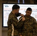 Two U.S. military personnel in MARPAT camouflage uniforms are conducting a Tactical Combat Casualty Care (TCCC) training demonstration. One service member is applying a pressure bandage or wound packing technique to the other's shoulder/upper arm area. A presentation slide from ASM TCCC is visible in the background, displaying instructions on wound packing and pressure bandage application.