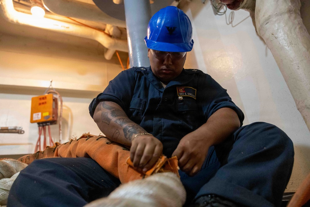 A U.S. Navy sailor identified by name tag as BM3 E. Gonzalez is seated on a deck working with rope or line equipment. The sailor is wearing a blue hard hat with a Navy anchor insignia, navy blue coveralls, safety glasses, and has visible tattoos on the right arm. The setting appears to be an interior compartment or lower deck area of a naval vessel.