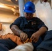 A U.S. Navy sailor identified by name tag as BM3 E. Gonzalez is seated on a deck working with rope or line equipment. The sailor is wearing a blue hard hat with a Navy anchor insignia, navy blue coveralls, safety glasses, and has visible tattoos on the right arm. The setting appears to be an interior compartment or lower deck area of a naval vessel.