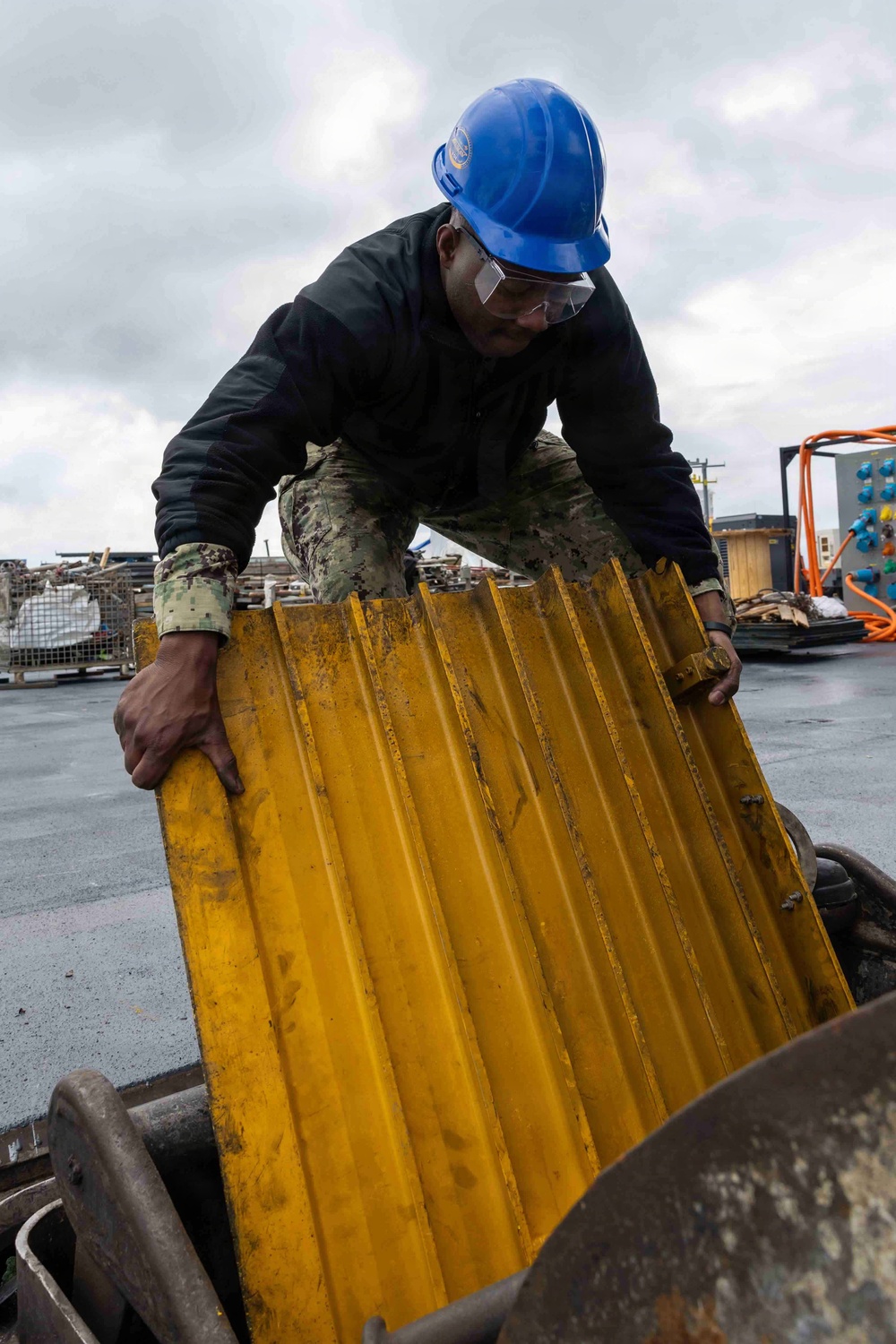 A worker wearing a blue hard hat, safety glasses, and camouflage military-style pants is handling a large yellow corrugated metal panel, appearing to load or move it into a metal container or bin. The setting appears to be an industrial or shipyard environment with various materials and equipment visible in the background. The overcast sky and wet-looking surfaces suggest outdoor work in damp conditions.