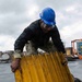 A worker wearing a blue hard hat, safety glasses, and camouflage military-style pants is handling a large yellow corrugated metal panel, appearing to load or move it into a metal container or bin. The setting appears to be an industrial or shipyard environment with various materials and equipment visible in the background. The overcast sky and wet-looking surfaces suggest outdoor work in damp conditions.