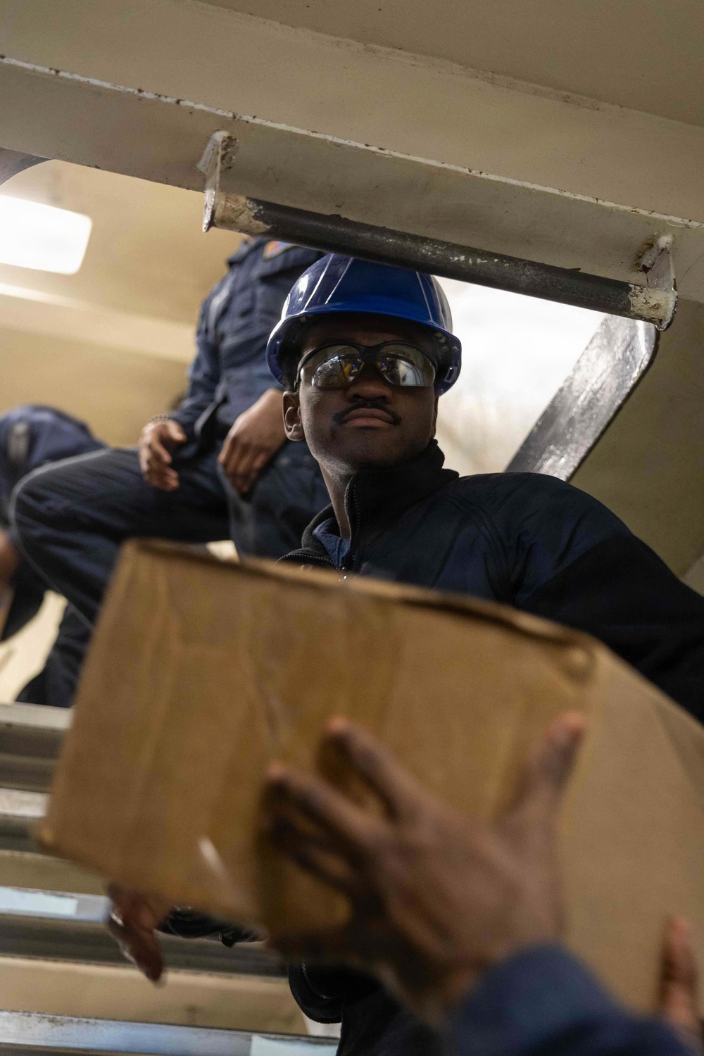 A worker wearing a blue hard hat and safety goggles is passing or receiving a cardboard box through a hatch or opening, likely aboard a ship or industrial vessel. The image is taken from a low angle looking upward, giving a dynamic perspective of the cargo transfer operation. At least one additional person in dark coveralls is visible in the background above the hatch.