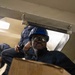 A worker wearing a blue hard hat and safety goggles is passing or receiving a cardboard box through a hatch or opening, likely aboard a ship or industrial vessel. The image is taken from a low angle looking upward, giving a dynamic perspective of the cargo transfer operation. At least one additional person in dark coveralls is visible in the background above the hatch.