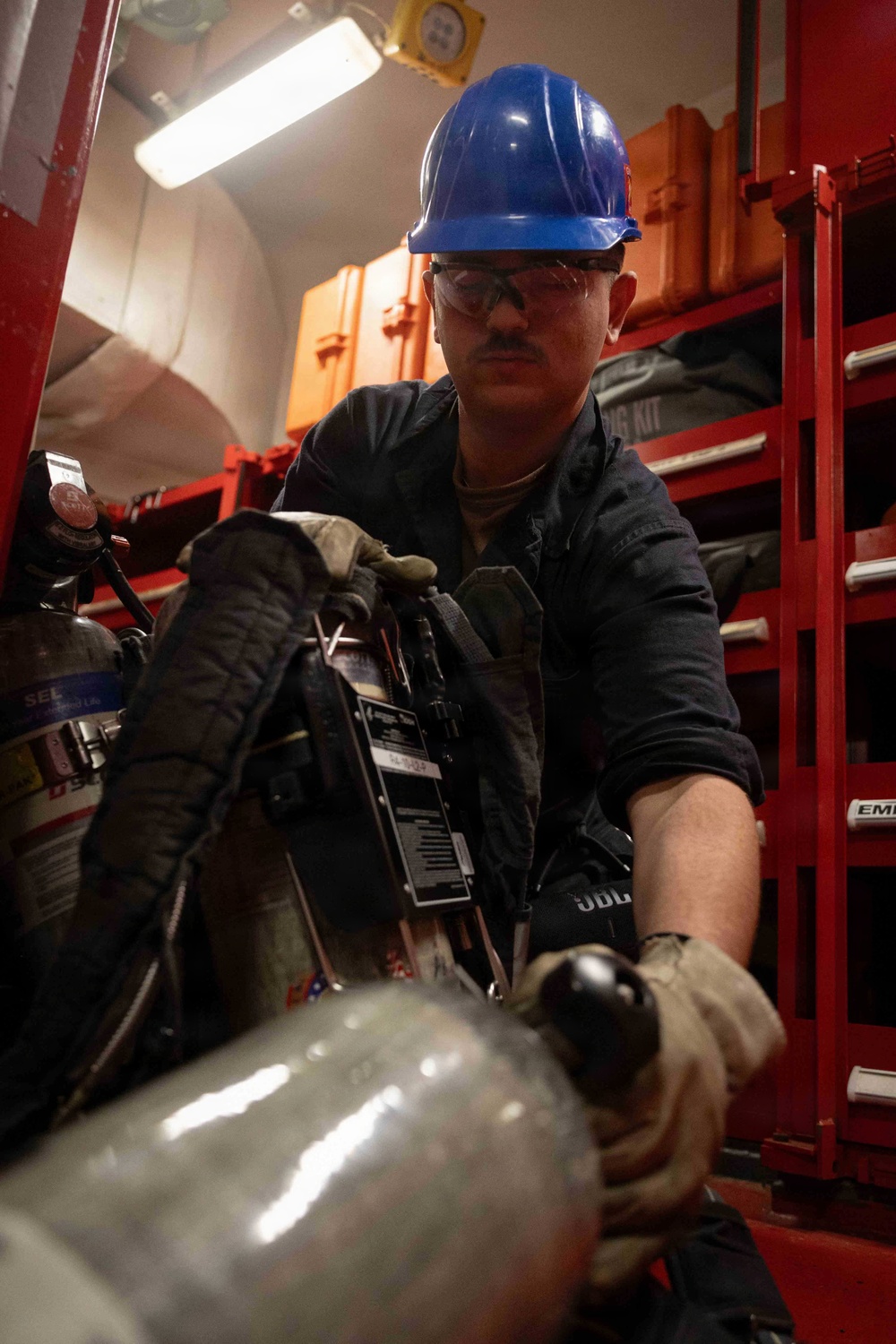 A worker wearing a blue hard hat and safety glasses is handling equipment in what appears to be a storage or maintenance area. The individual is wearing dark coveralls and work gloves while manipulating what appears to be breathing apparatus or compressed gas equipment. The setting features red storage shelving units stocked with various equipment and orange cases in the background.
