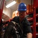 A worker wearing a blue hard hat and safety glasses is handling equipment in what appears to be a storage or maintenance area. The individual is wearing dark coveralls and work gloves while manipulating what appears to be breathing apparatus or compressed gas equipment. The setting features red storage shelving units stocked with various equipment and orange cases in the background.