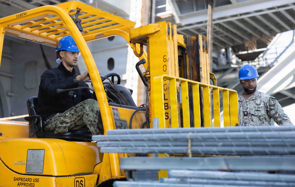 Two U.S. Navy personnel in camouflage uniforms and blue hard hats are operating a yellow forklift aboard a naval vessel. One sailor is driving the forklift while the other acts as a spotter/guide, overseeing the movement of metal materials. The forklift is labeled 'SHIPBOARD USE APPROVED' and bears the identifier '13-85741'.