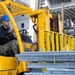 Two U.S. Navy personnel in camouflage uniforms and blue hard hats are operating a yellow forklift aboard a naval vessel. One sailor is driving the forklift while the other acts as a spotter/guide, overseeing the movement of metal materials. The forklift is labeled 'SHIPBOARD USE APPROVED' and bears the identifier '13-85741'.