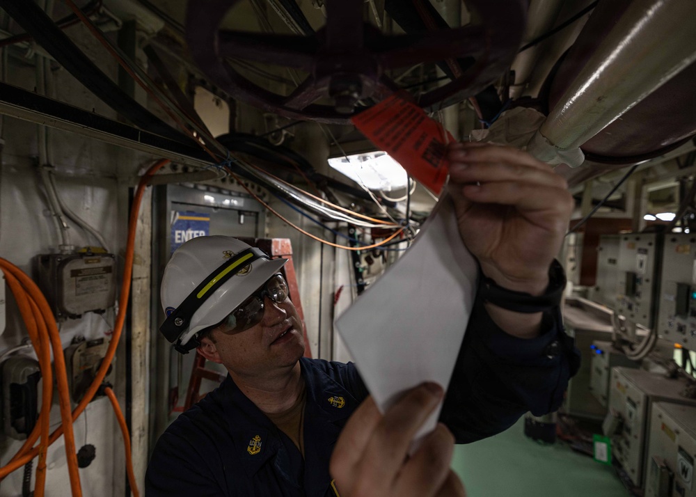 A U.S. Navy chief petty officer wearing a white hard hat and dark uniform is inspecting or attaching a red tag to overhead piping or cabling in what appears to be a ship's interior compartment. The individual is holding a white document or form in one hand while examining the red tag with the other. The environment shows a densely packed shipboard space with wiring, pipes, electrical panels, and equipment typical of a naval vessel.