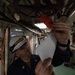 A U.S. Navy chief petty officer wearing a white hard hat and dark uniform is inspecting or attaching a red tag to overhead piping or cabling in what appears to be a ship's interior compartment. The individual is holding a white document or form in one hand while examining the red tag with the other. The environment shows a densely packed shipboard space with wiring, pipes, electrical panels, and equipment typical of a naval vessel.