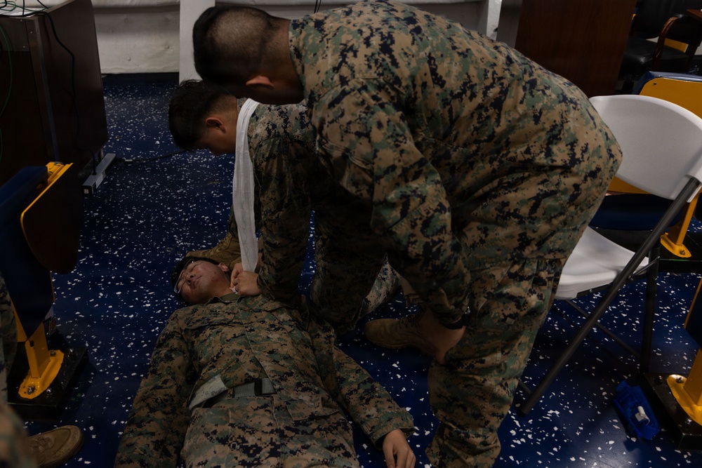 Two individuals in U.S. Marine Corps MARPAT camouflage uniforms are engaged in what appears to be a first aid or combat casualty care training exercise. One person is lying on a blue speckled floor while the other is leaning over them, appearing to apply or check a bandage or dressing near the head/neck area. The setting appears to be an indoor facility with folding chairs and equipment visible in the background.