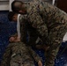 Two individuals in U.S. Marine Corps MARPAT camouflage uniforms are engaged in what appears to be a first aid or combat casualty care training exercise. One person is lying on a blue speckled floor while the other is leaning over them, appearing to apply or check a bandage or dressing near the head/neck area. The setting appears to be an indoor facility with folding chairs and equipment visible in the background.