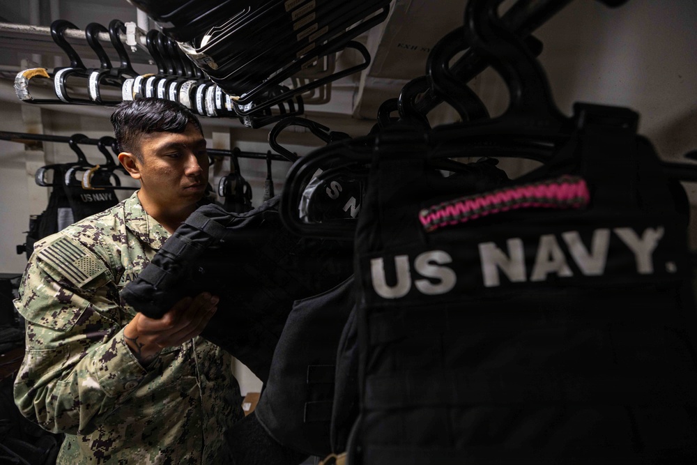 A U.S. Navy sailor in NWU Type III (digital camouflage) uniform is handling tactical gear, specifically what appears to be body armor or plate carriers, in a storage or armory room. Multiple black tactical vests labeled 'US NAVY' are hanging on racks in the background. The setting appears to be an organized equipment storage space aboard a naval vessel or installation.