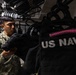 A U.S. Navy sailor in NWU Type III (digital camouflage) uniform is handling tactical gear, specifically what appears to be body armor or plate carriers, in a storage or armory room. Multiple black tactical vests labeled 'US NAVY' are hanging on racks in the background. The setting appears to be an organized equipment storage space aboard a naval vessel or installation.