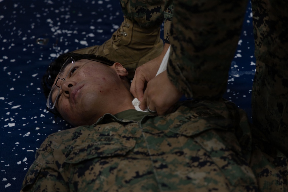 A military personnel in MARPAT (Marine Corps digital camouflage) uniform is lying on a blue mat while another service member administers what appears to be first aid or medical training. The person on the ground is wearing glasses pushed up on their forehead and is looking upward. This appears to be a combat casualty care or tactical combat casualty care (TCCC) training exercise.