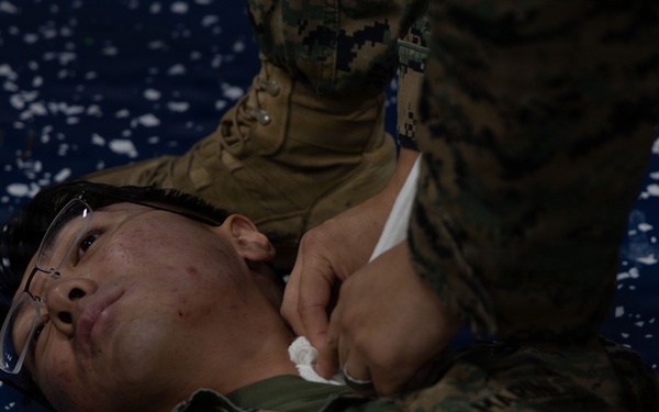 A military personnel in MARPAT (Marine Corps digital camouflage) uniform is lying on a blue mat while another service member administers what appears to be first aid or medical training. The person on the ground is wearing glasses pushed up on their forehead and is looking upward. This appears to be a combat casualty care or tactical combat casualty care (TCCC) training exercise.