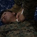 A military personnel in MARPAT (Marine Corps digital camouflage) uniform is lying on a blue mat while another service member administers what appears to be first aid or medical training. The person on the ground is wearing glasses pushed up on their forehead and is looking upward. This appears to be a combat casualty care or tactical combat casualty care (TCCC) training exercise.