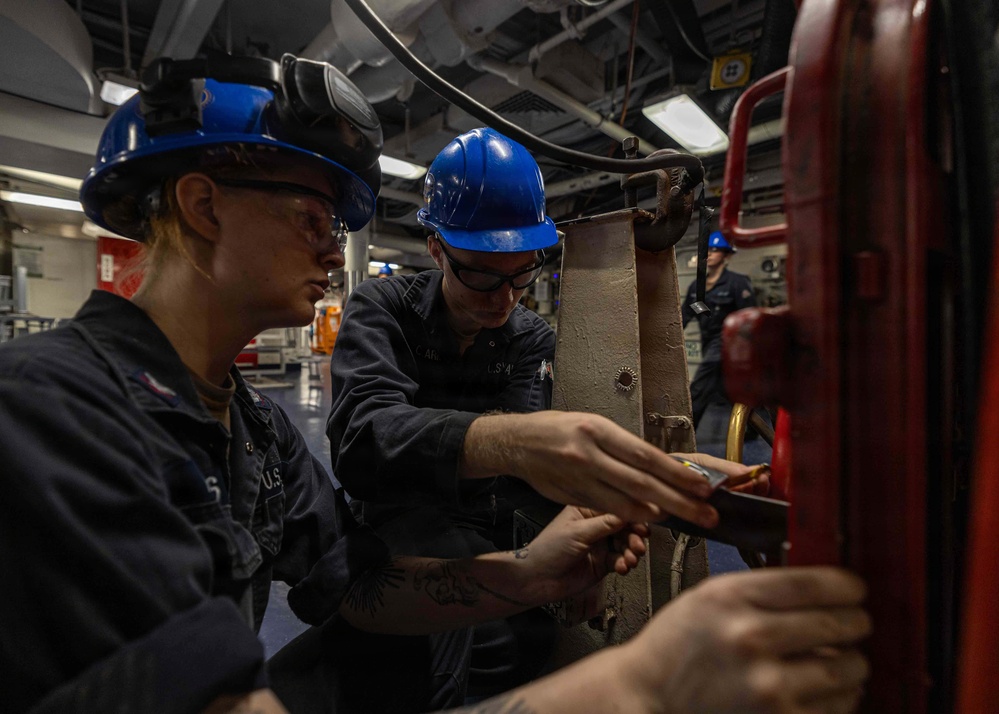 Two U.S. Navy sailors wearing blue hard hats and safety glasses are performing maintenance or repair work on mechanical equipment aboard what appears to be a naval vessel. They are working closely together on a red-painted piece of machinery or structural component. A third sailor in a blue hard hat is visible in the background observing or performing separate tasks.
