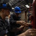 Two U.S. Navy sailors wearing blue hard hats and safety glasses are performing maintenance or repair work on mechanical equipment aboard what appears to be a naval vessel. They are working closely together on a red-painted piece of machinery or structural component. A third sailor in a blue hard hat is visible in the background observing or performing separate tasks.
