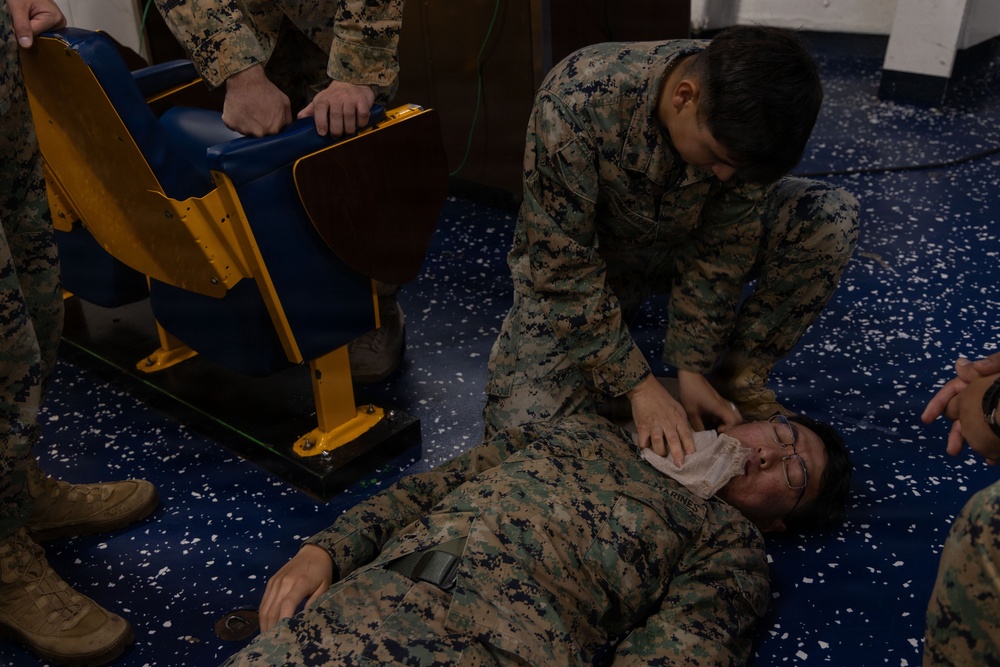 The image shows U.S. Marines in MARPAT camouflage uniforms conducting what appears to be a first aid or combat casualty care training exercise. One Marine is lying on the floor while another kneels over them, applying what appears to be a bandage or dressing to the face/head area. The setting appears to be an indoor facility with blue speckled flooring and stadium-style seating visible.