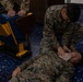 The image shows U.S. Marines in MARPAT camouflage uniforms conducting what appears to be a first aid or combat casualty care training exercise. One Marine is lying on the floor while another kneels over them, applying what appears to be a bandage or dressing to the face/head area. The setting appears to be an indoor facility with blue speckled flooring and stadium-style seating visible.