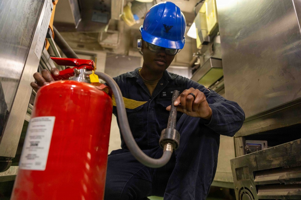 A military or naval personnel member wearing a blue hard hat and safety glasses is inspecting or maintaining a red fire extinguisher in what appears to be a ship's interior compartment. The individual is dressed in a dark navy coverall uniform with visible rank/name patches and is handling the extinguisher's hose nozzle. The setting suggests a routine safety inspection or maintenance task aboard a naval vessel.