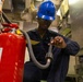 A military or naval personnel member wearing a blue hard hat and safety glasses is inspecting or maintaining a red fire extinguisher in what appears to be a ship's interior compartment. The individual is dressed in a dark navy coverall uniform with visible rank/name patches and is handling the extinguisher's hose nozzle. The setting suggests a routine safety inspection or maintenance task aboard a naval vessel.