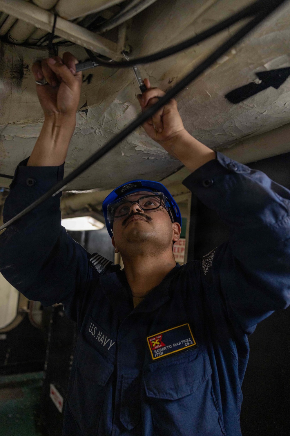 A U.S. Navy sailor identified as Roberto Martinez, CS-3, is performing maintenance work overhead, using tools on cables or conduits attached to a ship's ceiling. He is wearing standard Navy working uniform (NWU), a blue hard hat, and safety glasses. The setting appears to be an interior compartment of a naval vessel.